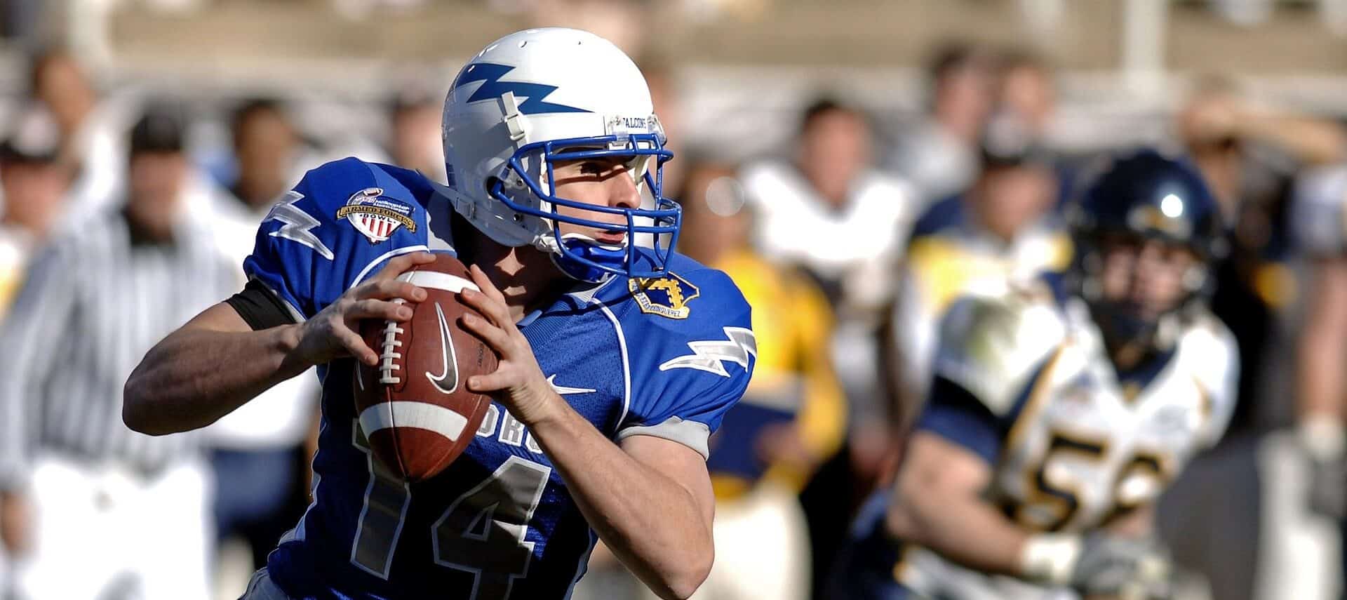 Image of a foorball quarterback holding the ball and wearing a blue jersey and white helmet with a lightening bolt.