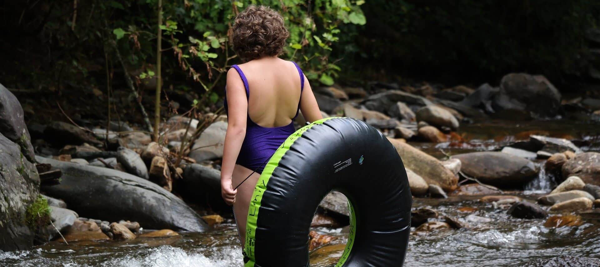 A woman wearing a purple swim suit standing in a river with an inner tube leaning on her.
